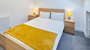 A hotel room with a bed, two bedside tables with lamps, a radiator, and a cityscape painting on the wall. - Jackson Court Apartment 3 (Redcar)