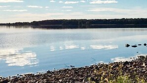 Un lac paisible, bordé d’une rive rocheuse, avec une ligne d’arbres qui se dessine à l’horizon sous un ciel dégagé.