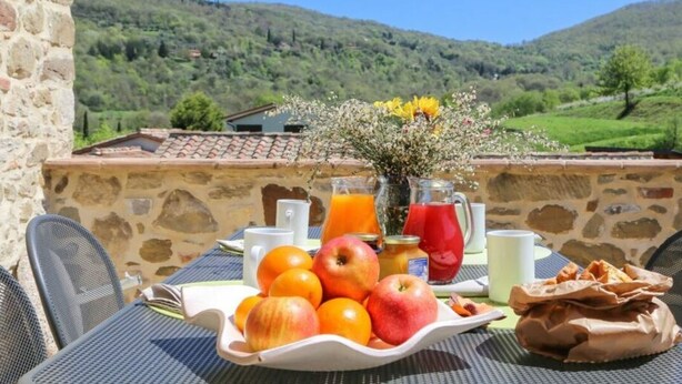 Ein Tisch mit Obst, Getränken und einer Tüte Snacks auf einem Balkon mit einer Steinmauer und Blick auf grüne Hügel.