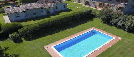 A large rectangular swimming pool surrounded by well-maintained grass, with a stone building and a smaller house in the background.