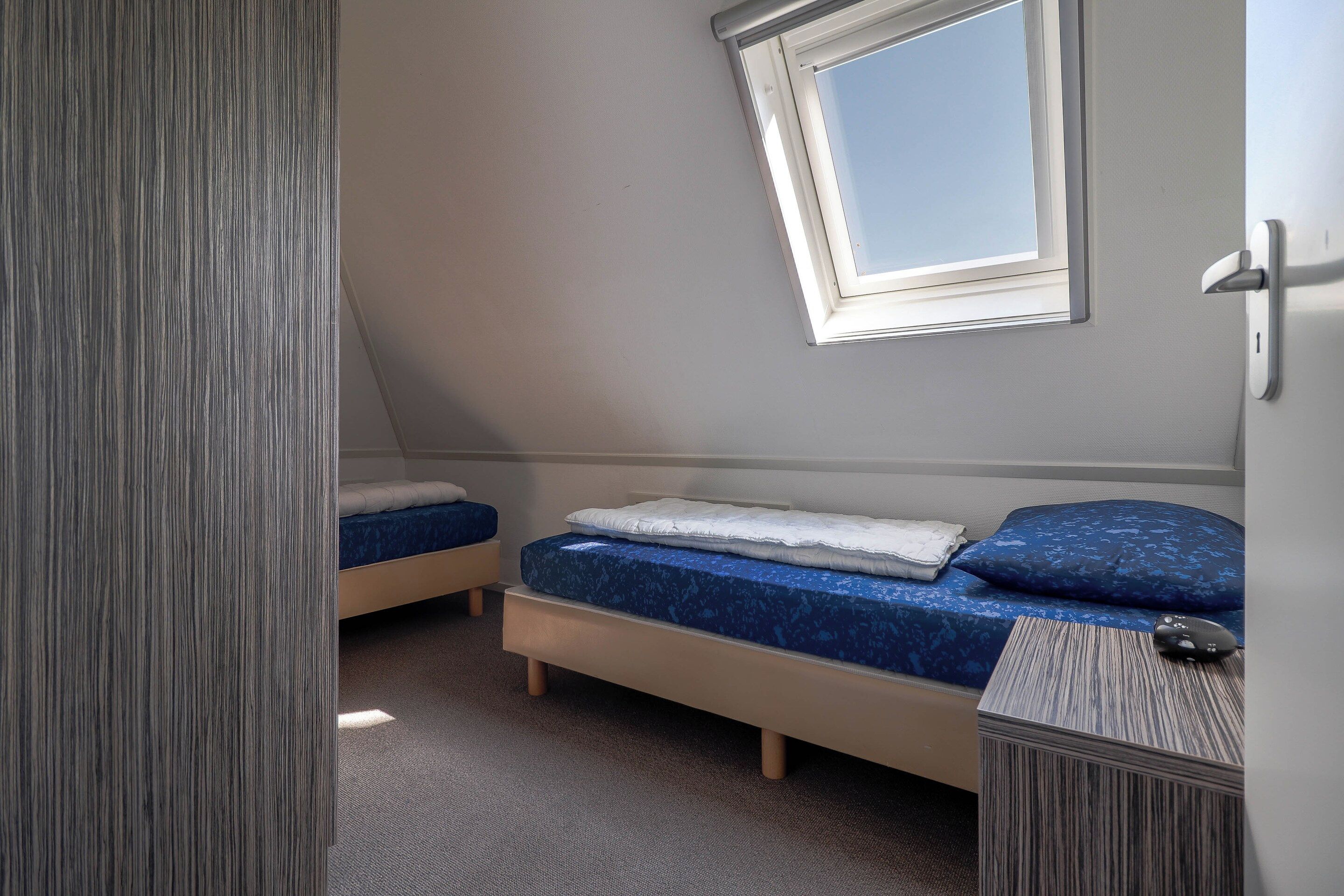A small, single-room accommodation with a sloped ceiling, a window, a bed, a bedside table, and a door.