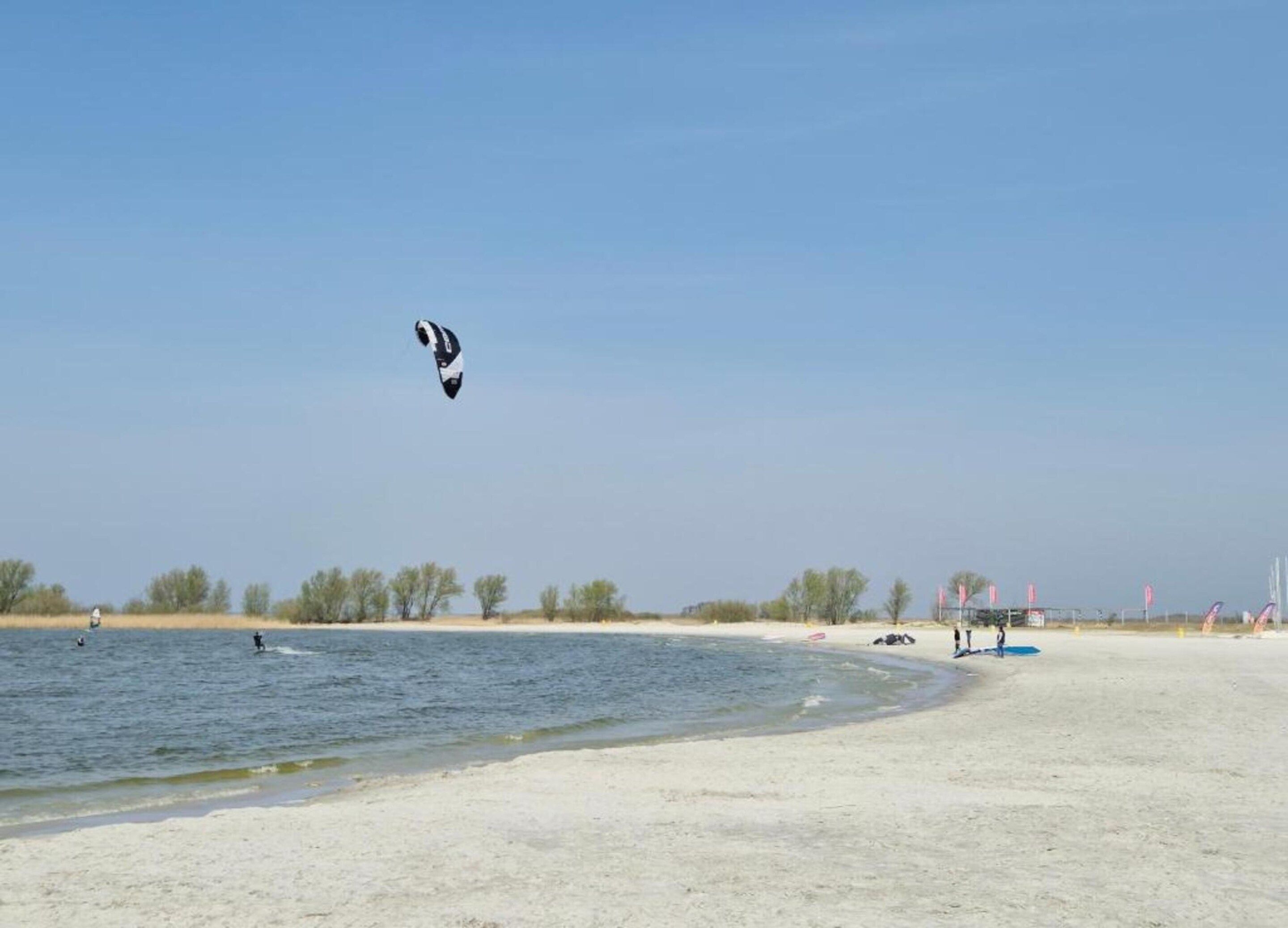 Une plage avec un ciel dégagé, une personne pratiquant le kitesurf et quelques personnes sur le sable.