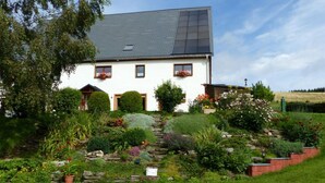 A white house with a grey roof, surrounded by a well-maintained garden with various plants and flowers. - Holiday Apartment Family Kreissl (Oberwiesenthal)