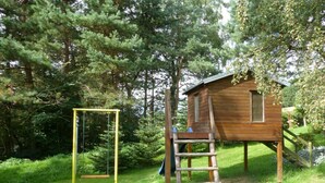 A wooden playhouse with a ladder and a swing set in a grassy area surrounded by trees. - Holiday Apartment Family Kreissl (Oberwiesenthal)