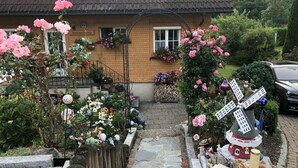 A house with a stone pathway leading to a wooden gate, surrounded by a garden with various flowers and a windmill decoration.