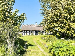 Une maison traditionnelle à toit de chaume, accessible par un étroit sentier, entourée d’une végétation luxuriante et d’arbres.