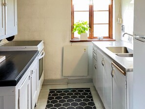 Une cuisine avec des meubles blancs, un plan de travail noir, un petit réfrigérateur, une fenêtre avec une plante et un pare-feu en carreaux.