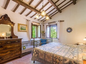 A bedroom with a wooden headboard, a large bed with a quilt, a dresser with drawers, a mirror, and two windows with curtains. - Belvilla by OYO Villa Dipinti (Serra San Quirico)