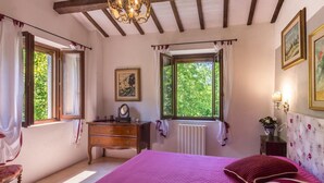 A bedroom with a large bed, wooden dresser, two windows, a radiator, and a chandelier. - Belvilla by OYO Villa Dipinti (Serra San Quirico)
