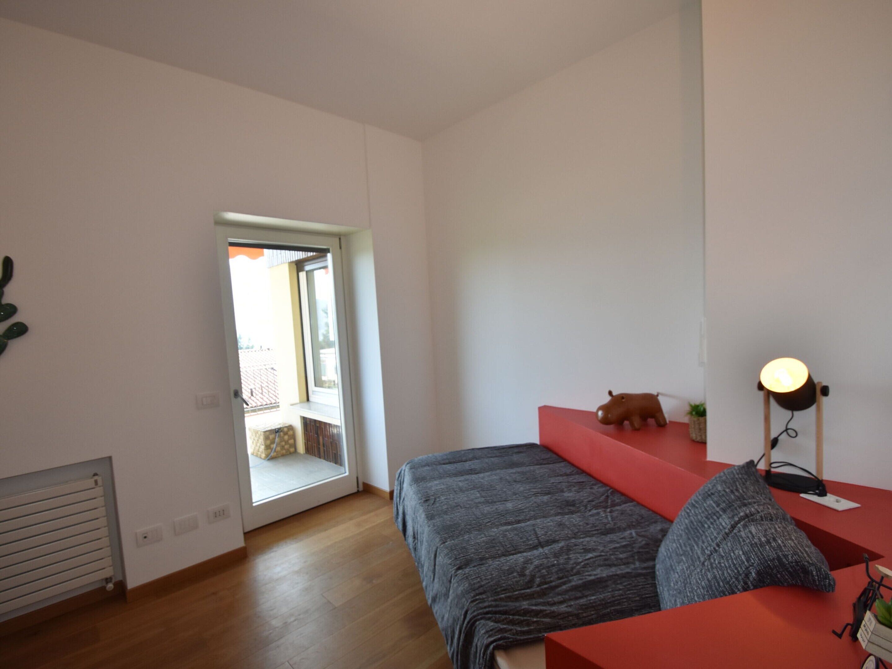 Une chambre à coucher avec un lit, une table de chevet rouge, une lampe, une petite plante et une vue sur l’extérieur, visible à travers une porte.