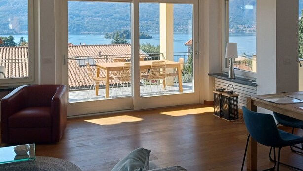 A spacious room with a wooden floor, a red chair, a grey sofa, and a dining table with chairs. Large windows offer a view of a balcony with a table and chairs, overlooking a lake and mountains. - Apartment in a Prime Location on Lake Maggiore (Verbania)