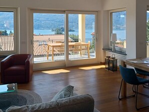 A spacious room with a wooden floor, a red chair, a grey sofa, and a dining table with chairs. Large windows offer a view of a balcony with a table and chairs, overlooking a lake and mountains. - Apartment in a Prime Location on Lake Maggiore (Verbania)