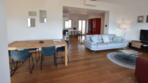 A modern living room with a wooden dining table, two blue chairs, a grey sofa with cushions, a red armchair, a glass coffee table, and a flat-screen TV. - Apartment in a Prime Location on Lake Maggiore (Verbania)