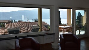A modern living room with a large window offering a view of a lake and mountains. There is a dining area with a table and chairs visible through the window. - Apartment in a Prime Location on Lake Maggiore (Verbania)