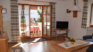 Un salon chaleureux avec un plancher en bois, un téléviseur, un coin repas avec une table et des chaises, et une porte-fenêtre coulissante donnant sur une terrasse extérieure.