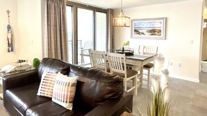 Una sala de estar con un sofá de cuero negro, una mesa de comedor con sillas y un ventanal con cortinas.