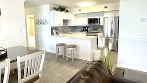 Una cocina moderna con armarios blancos, una isla central con taburetes, electrodomésticos de acero inoxidable y un comedor con sofá de cuero marrón y sillas blancas.