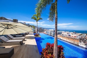 A rooftop pool with lounge chairs and umbrellas, surrounded by palm trees and a view of the sea and buildings. - New Downtown Condo w/ Private Dipping Pool (Puerto Vallarta)