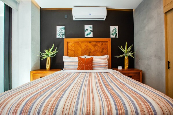 A bed with a striped bedspread, two nightstands with potted plants, a wooden headboard, and wall art. - New Downtown Condo w/ Private Dipping Pool (Puerto Vallarta)