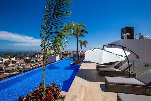 A rooftop infinity pool with lounge chairs and palm trees, overlooking a cityscape.