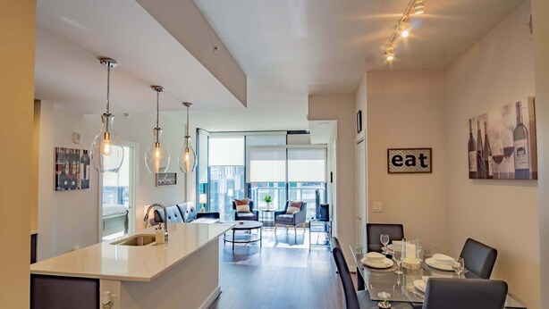 A modern kitchen with a central island, dining area with a glass table, and a living space with a sofa and chairs.