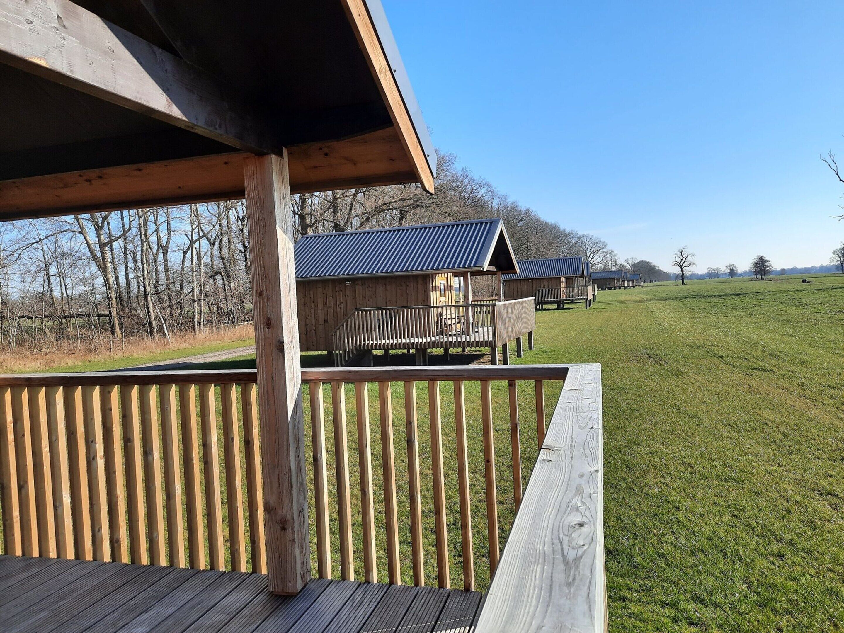 Une terrasse en bois avec une rambarde, un espace abrité et une vue sur un bâtiment doté d’un toit en métal, situé dans un champ verdoyant.