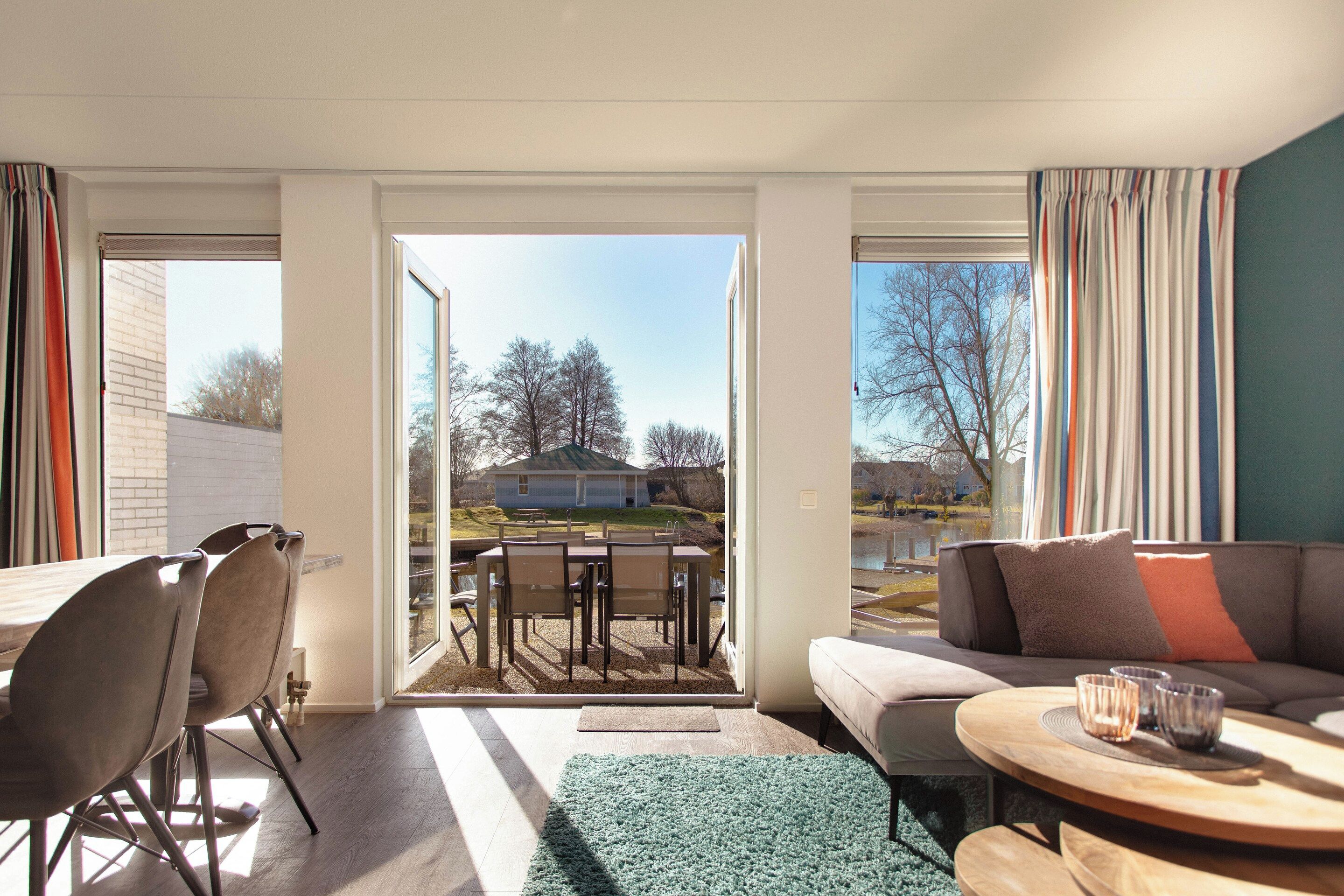 A modern living room with a sofa, chairs, a dining table, and a view of a house and trees through large windows.