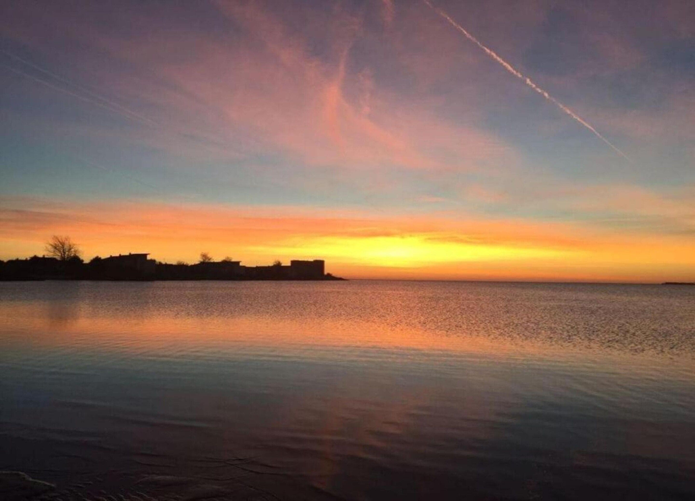 A calm body of water reflecting a sunset sky with a visible contrail.