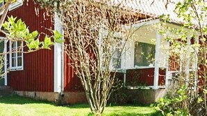 A red building with a white porch, surrounded by a grassy yard and a tree with bare branches. - 5 Person Holiday Home in Gamleby (Gamleby)