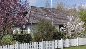 A house with a white picket fence, surrounded by greenery and flowering trees. - Long Comfortable Holiday Residence (Süderlügum)