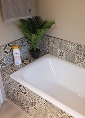 A bathroom with a white bathtub, a potted plant, and a bottle of Elkos body lotion.