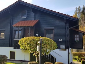 A two-story wooden house with a dark facade, a red-tiled roof, and a small garden with a neatly trimmed bush.