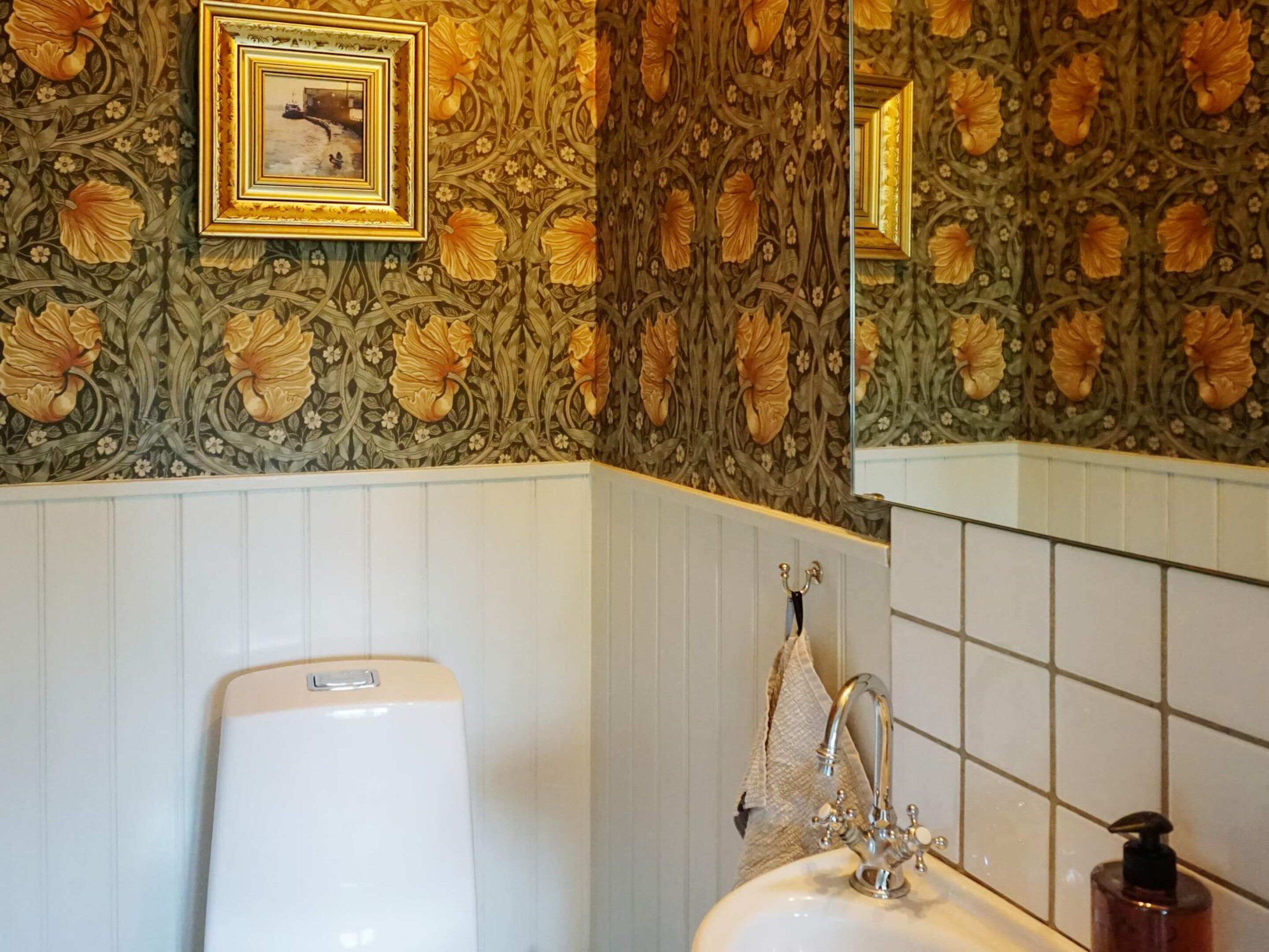 Une salle de bain avec un lavabo blanc, un miroir et un mur recouvert d’un papier peint à motifs floraux.