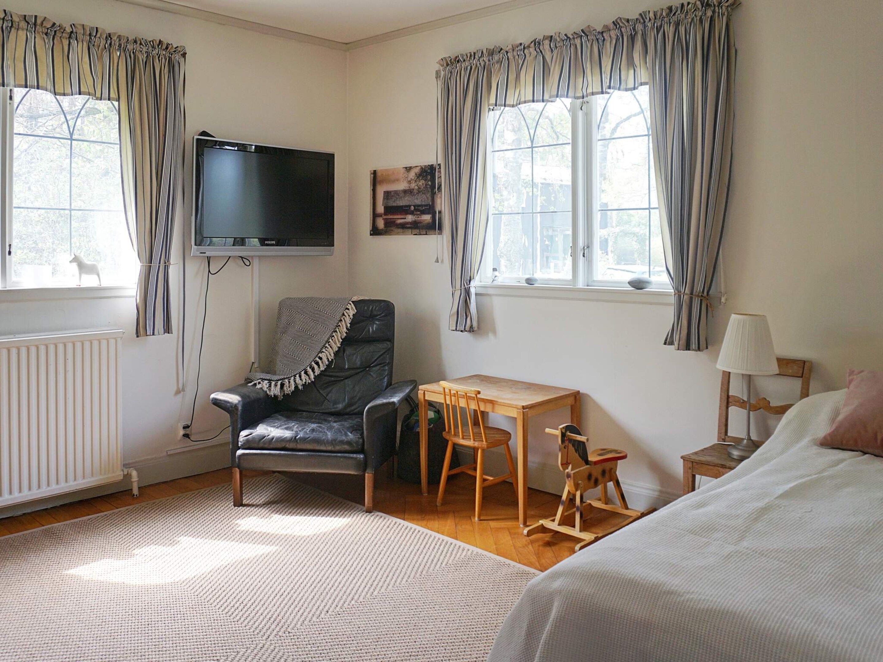 A room with a black leather chair, a wooden table, a television mounted on the wall, and a bed with a white blanket.