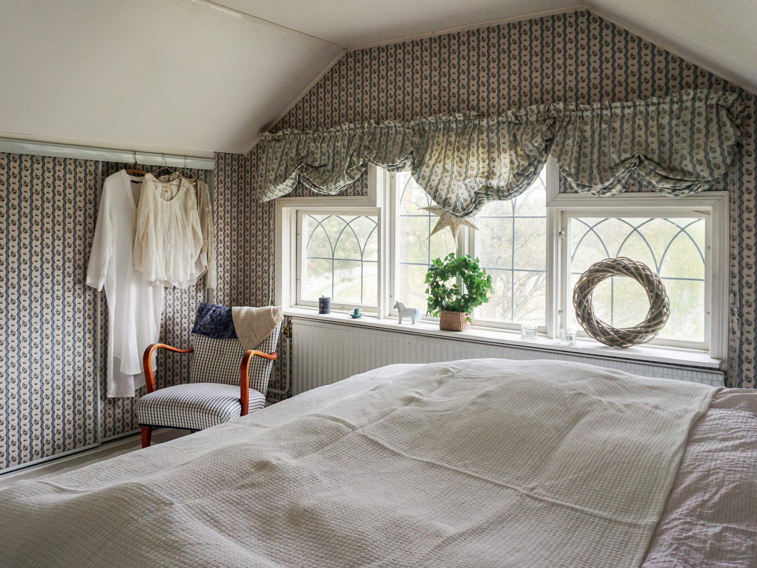 A bedroom with a large bed, a chair, a window with curtains, and a wall with patterned wallpaper.