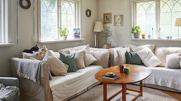 A cozy living room with a sofa, a coffee table, and a lamp. There are windows with curtains and a view of greenery outside.