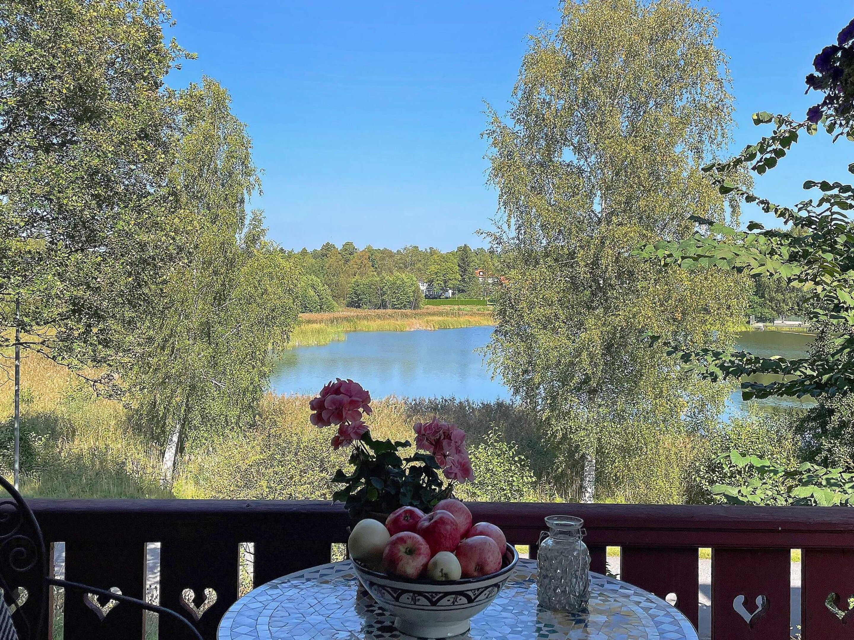 A table with a bowl of fruit and flowers, overlooking a lake with trees and a house in the background.