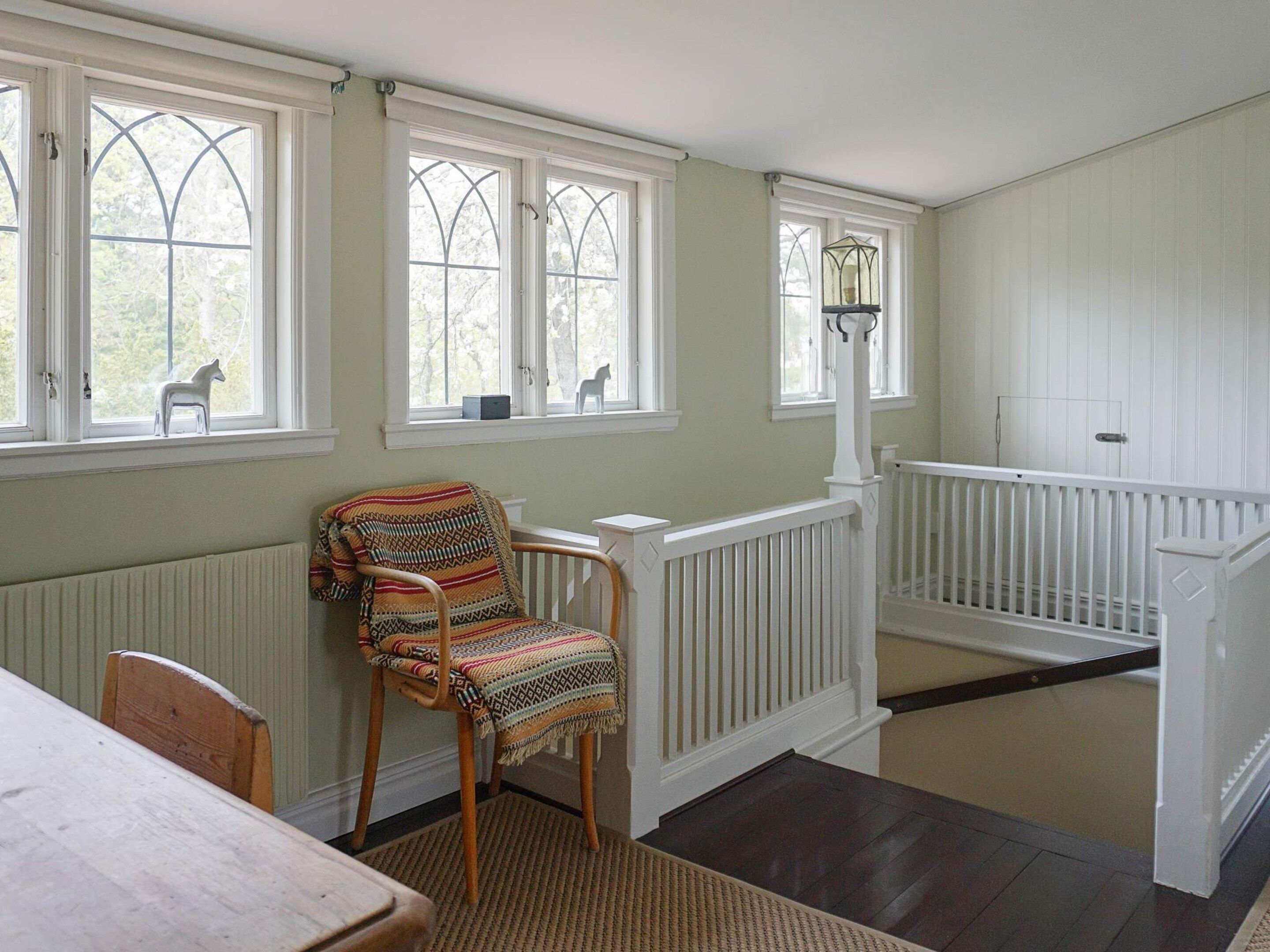 A room with a wooden chair, a table, a white railing, and a window with a view of trees.