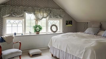Quarto com cama grande, uma cadeira, um banquinho e uma janela decorada com uma guirlanda, além de uma planta.