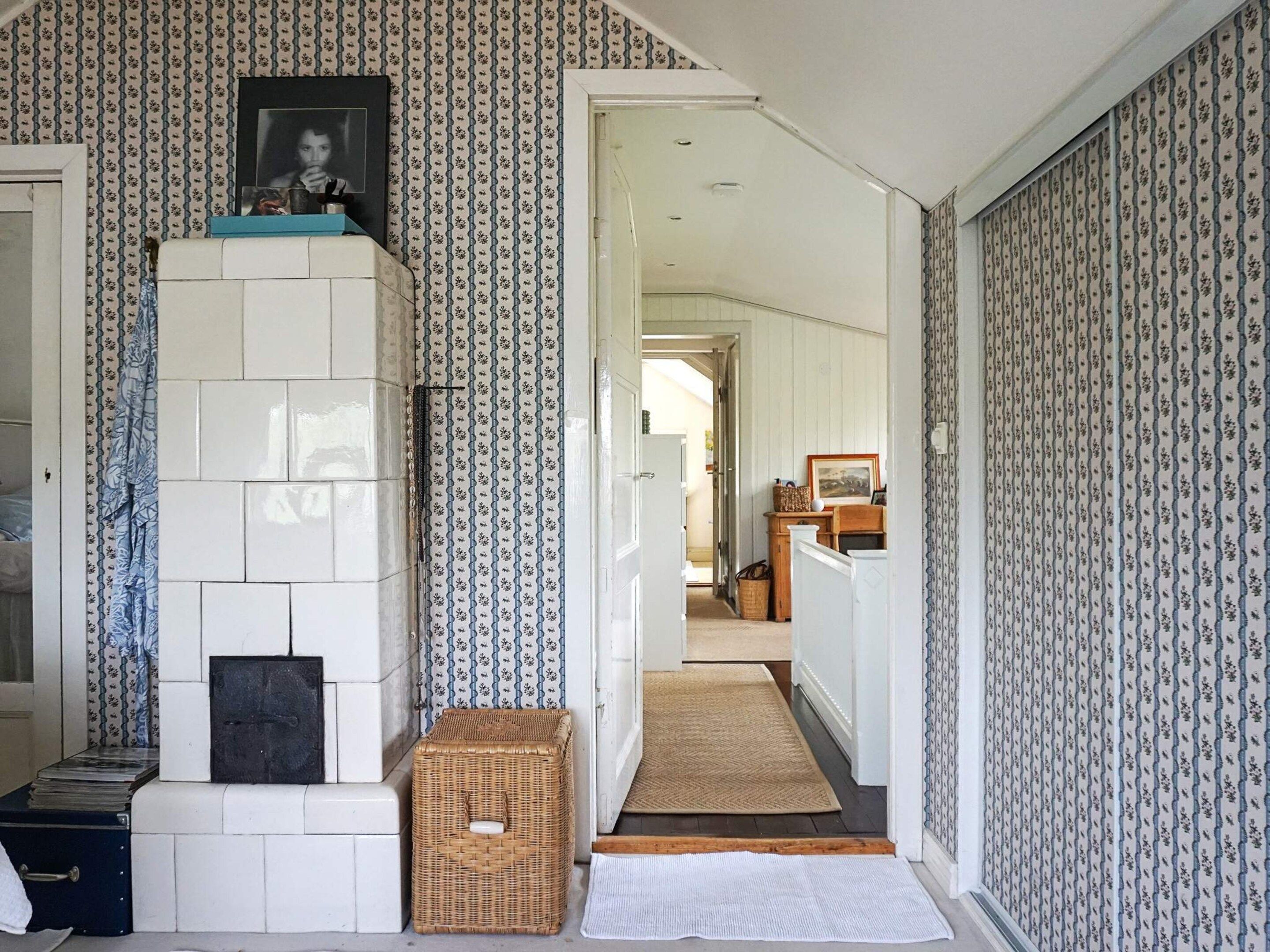 A hallway with patterned wallpaper, a fireplace, a wicker basket, and a rug leading to another room.