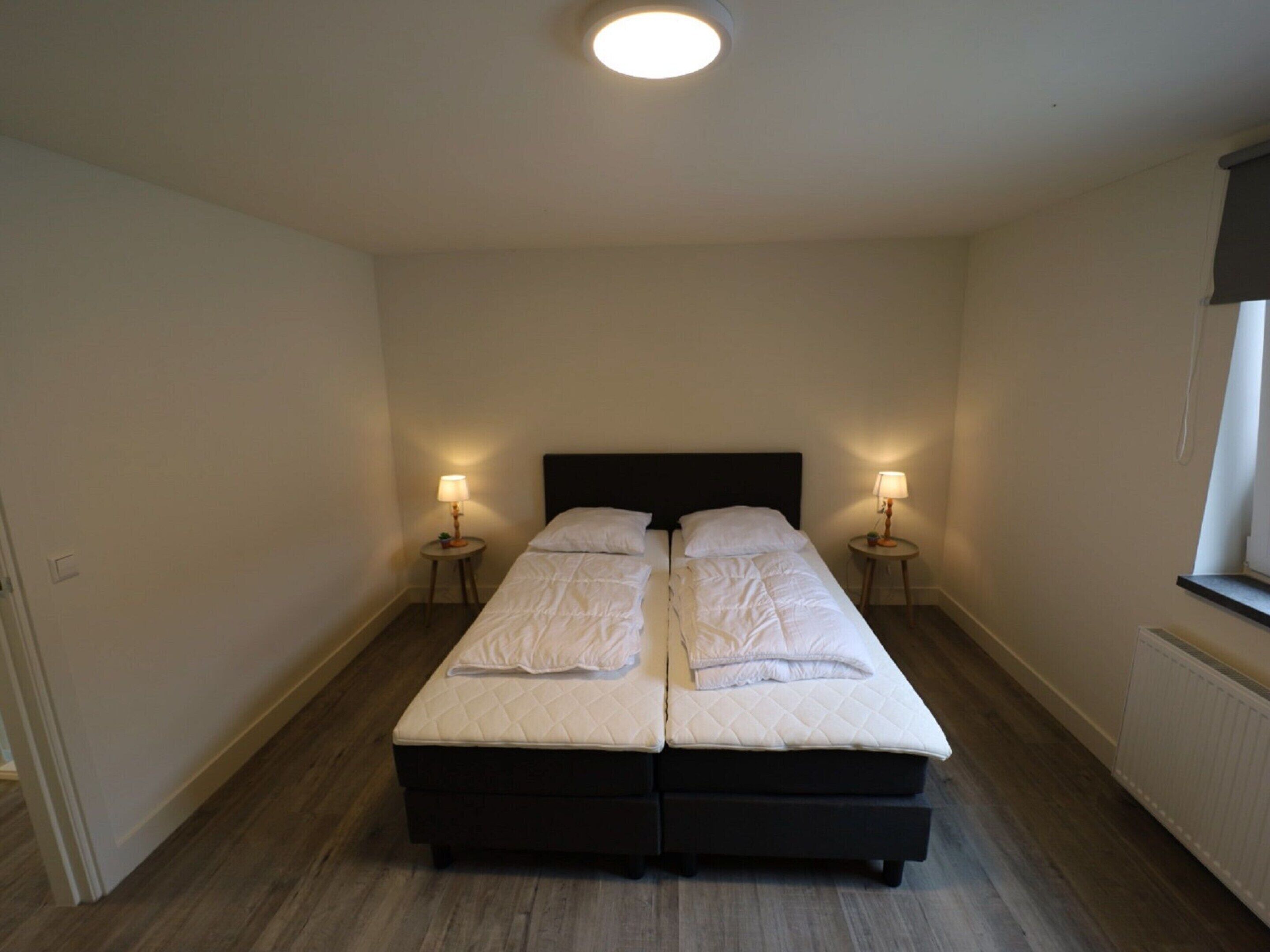 Una cama doble con colchón acolchado y dos mesitas de noche con lámparas, en una habitación con suelo de madera y pared lisa.
