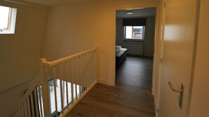 A narrow hallway with wooden flooring, a white railing, and a door leading to a bedroom with a window and a radiator. - Detached Holiday Home Near Alkmaar (Hensbroek)