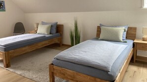 A room with two wooden beds, each with a grey mattress and matching pillows. There is a desk with a lamp, a chair, and a tall green plant. - Holiday Home Heinrich (Wurster Nordseeküste)
