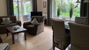 A living room with a sofa, armchair, and dining table. There is a TV and a window with a view of greenery. - Holiday Home Heinrich (Wurster Nordseeküste)
