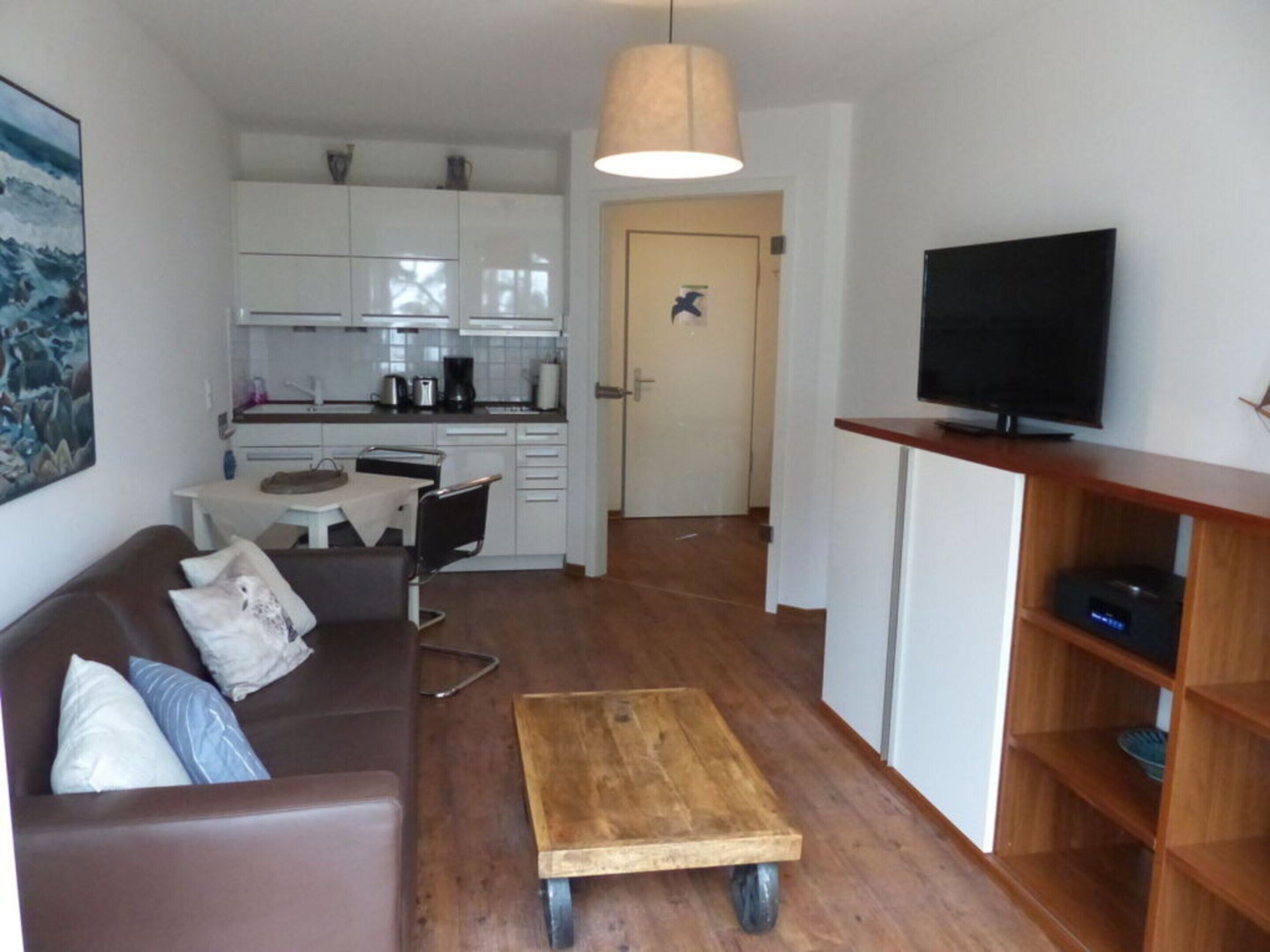Un espace de vie compact comprenant un coin cuisine, une petite table basse en bois sur roulettes, un canapé marron avec des coussins et un téléviseur posé sur un meuble en bois.