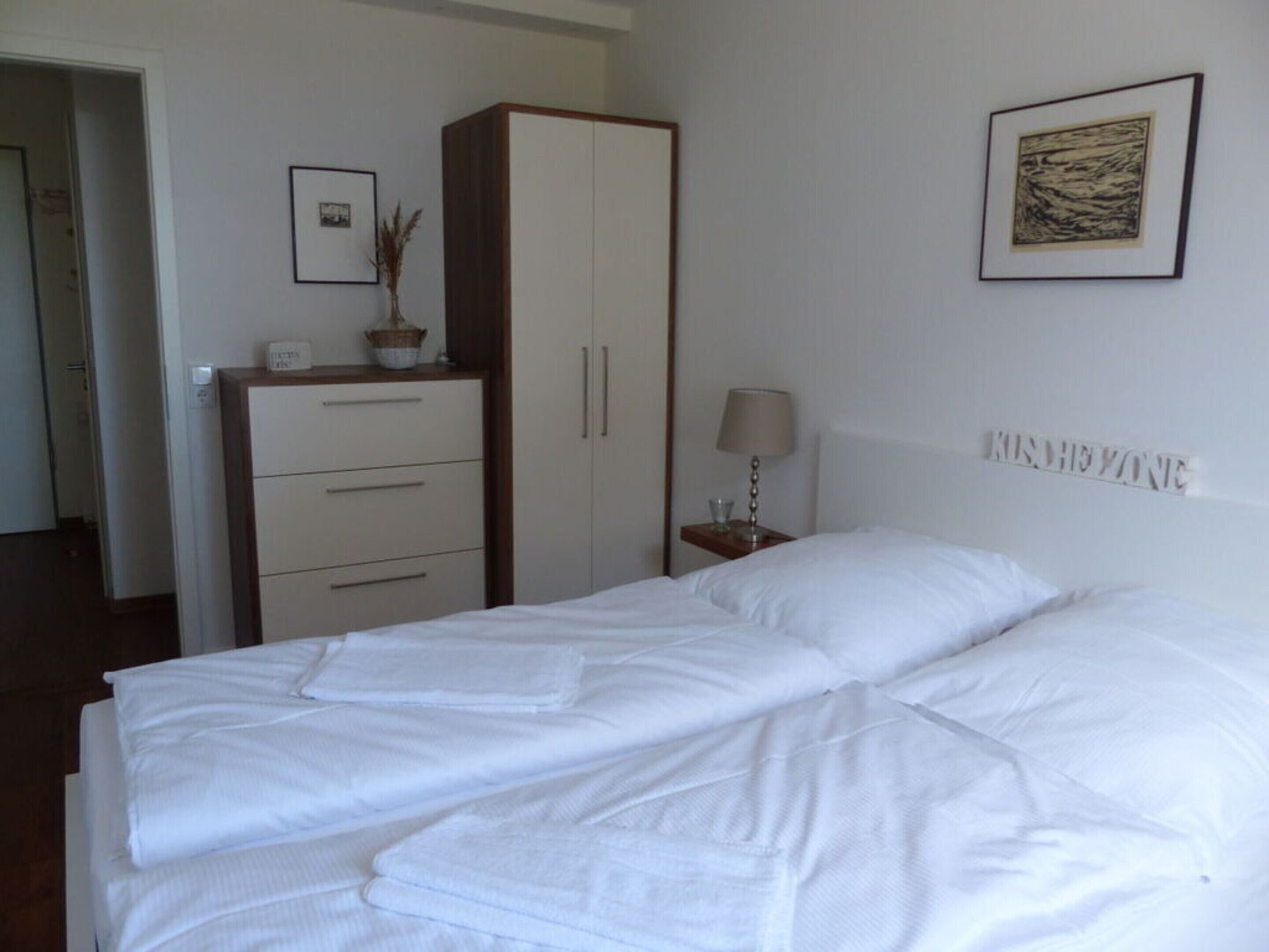 Un lit bien fait, avec du linge de lit blanc, une table de chevet avec une lampe, un petit tableau encadré et une commode avec des tiroirs.