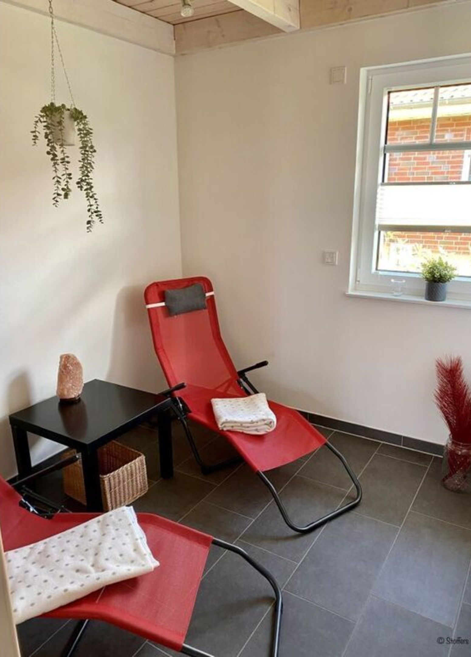 A small, clean room with a red chair, a small table, and a window with a view of a brick building.