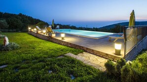 Una piscina con lettini e luci, circondata da un solarium in pietra e erba, con vista sulle colline e una luna nel cielo.