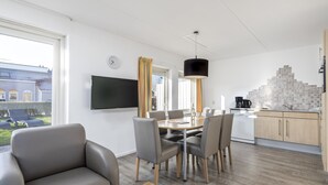 A modern living room with a sofa, dining table, and chairs, a kitchen with a tiled backsplash, and a large window with curtains. - Bungalow in Renesse Near North Sea Beach (Renesse)