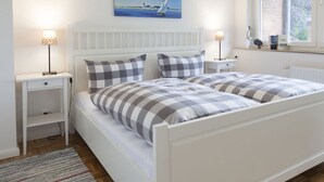 Quarto com cama branca, almofadas xadrez, mesa de cabeceira com abajur, pintura de um veleiro e janela com vista para árvores.
